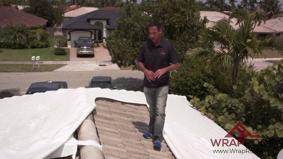 Tile roof wrapped and dry under white Wraproof membrane while a neighboring roof is still under a blue tarp